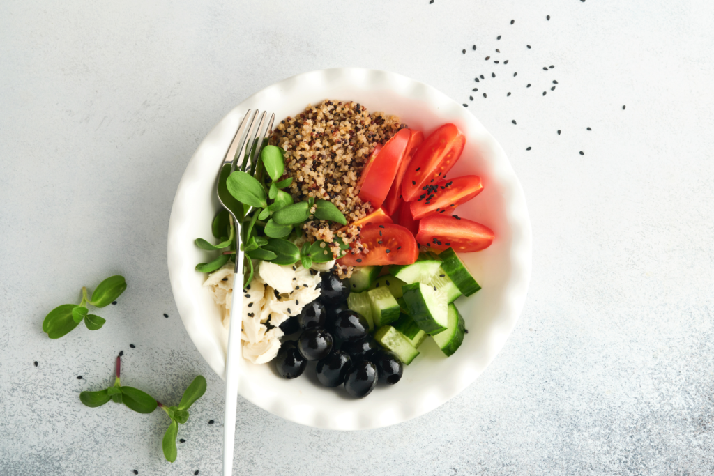 A healthy balanced meal with quinoa, tomatoes, cucumber, olives, leafy greens and lean protein served in a white bowl.