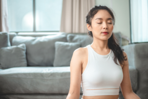A woman sitting indoors with her eyes closed while practicing mindfulness meditation and deep breathing for relaxation.
