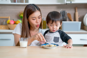 Mother helping her young child eat a meal at the dining table.