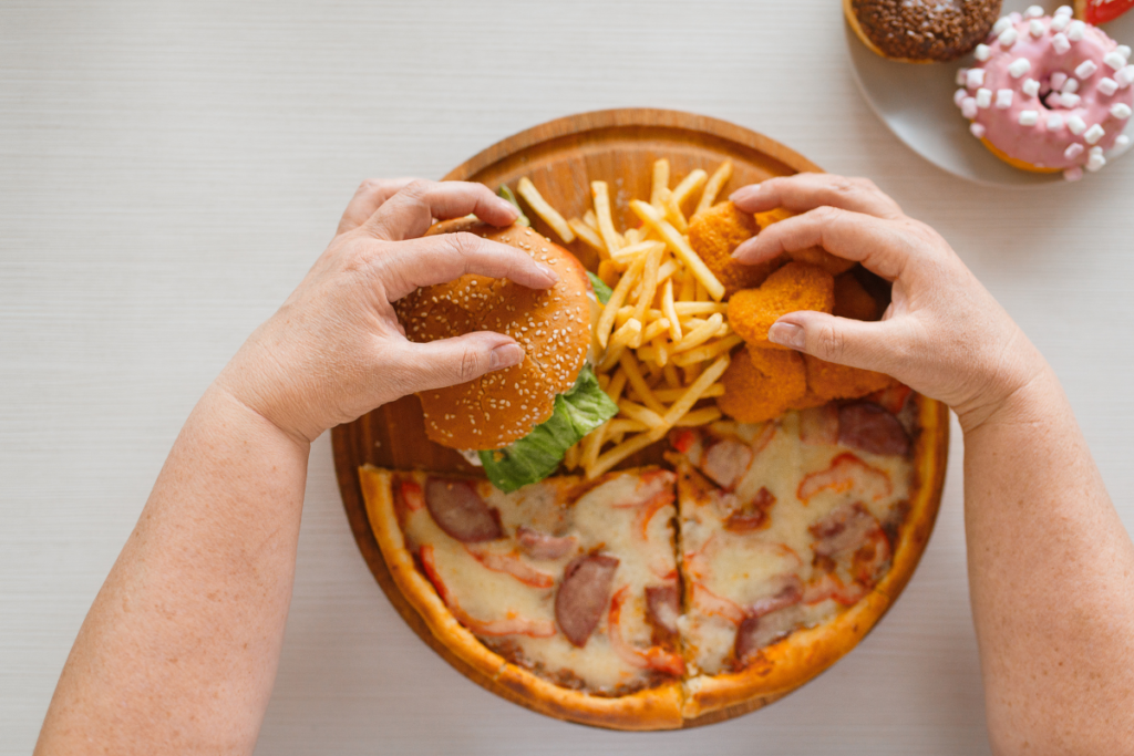 Person eating a burger, fries and pizza with doughnuts nearby.